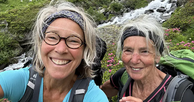 Twee oudere vrouwen met rugzakken glimlachen voor een selfie terwijl ze wandelen langs een stromend beekje in een weelderig, groen berggebied met bloeiende roze bloemen.
